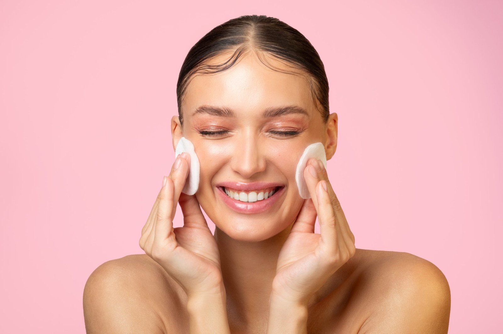 daily skincare. excited young lady cleaning face with cotton pads and smiling, doing skin care routine over pink background
